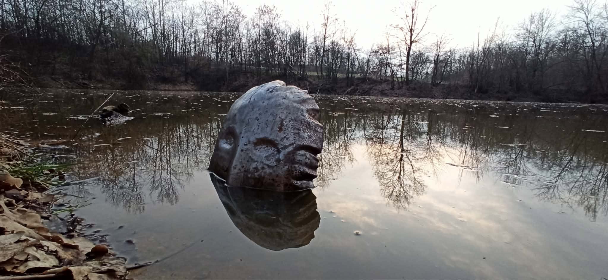 Volto scolpito in ferro da Diego Donadei, riflesso nell’acqua tra gli alberi